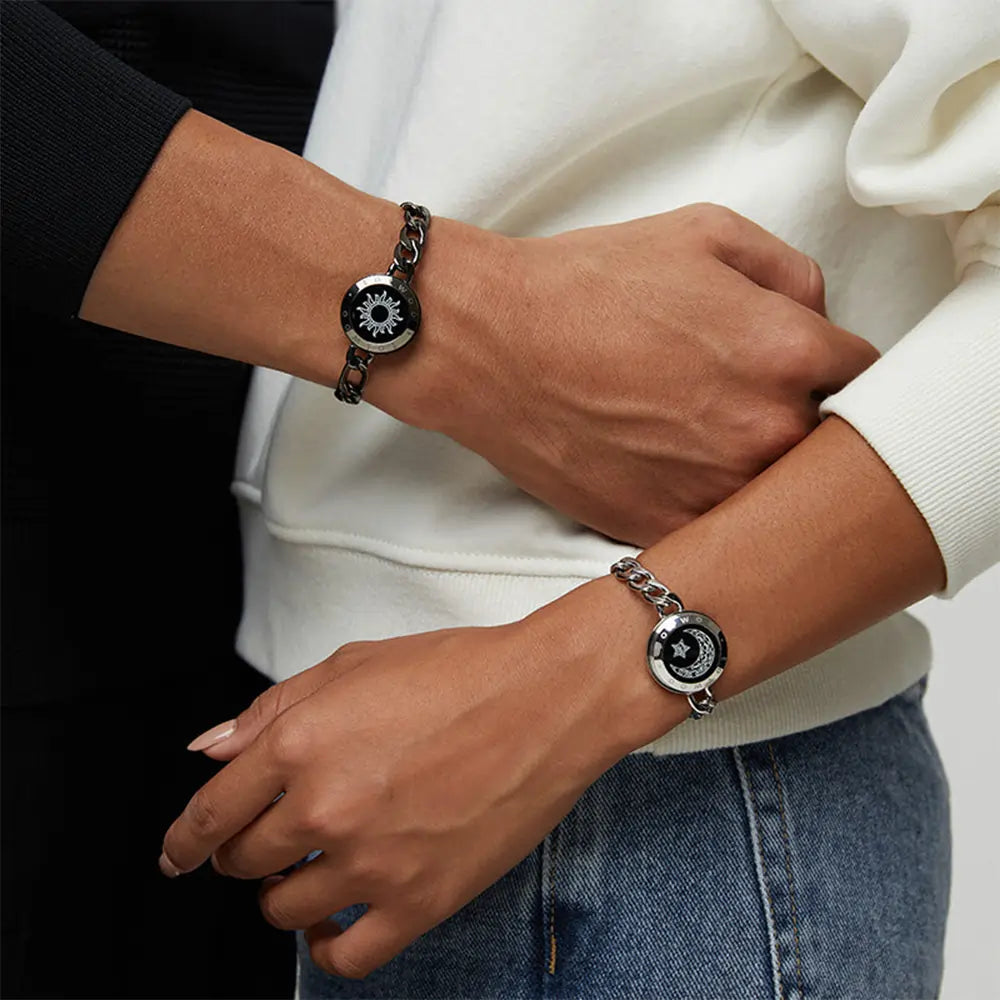 A close-up of two people wearing matching EntangleTies™ Sun & Moon Totwoo™ Bracelets. The bracelets feature a black chain and sun/moon designs on the wrists of both individuals.